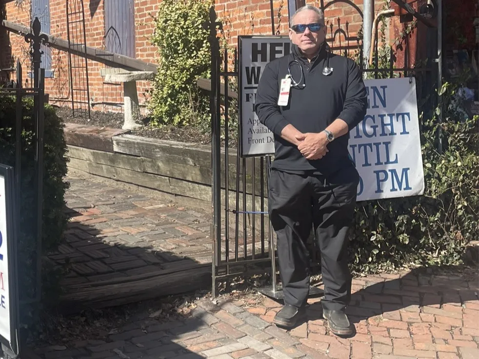 Dr Leve in clinic blacks with a stethoscope, standing at the clinic’s front gate beside a hiring notice.